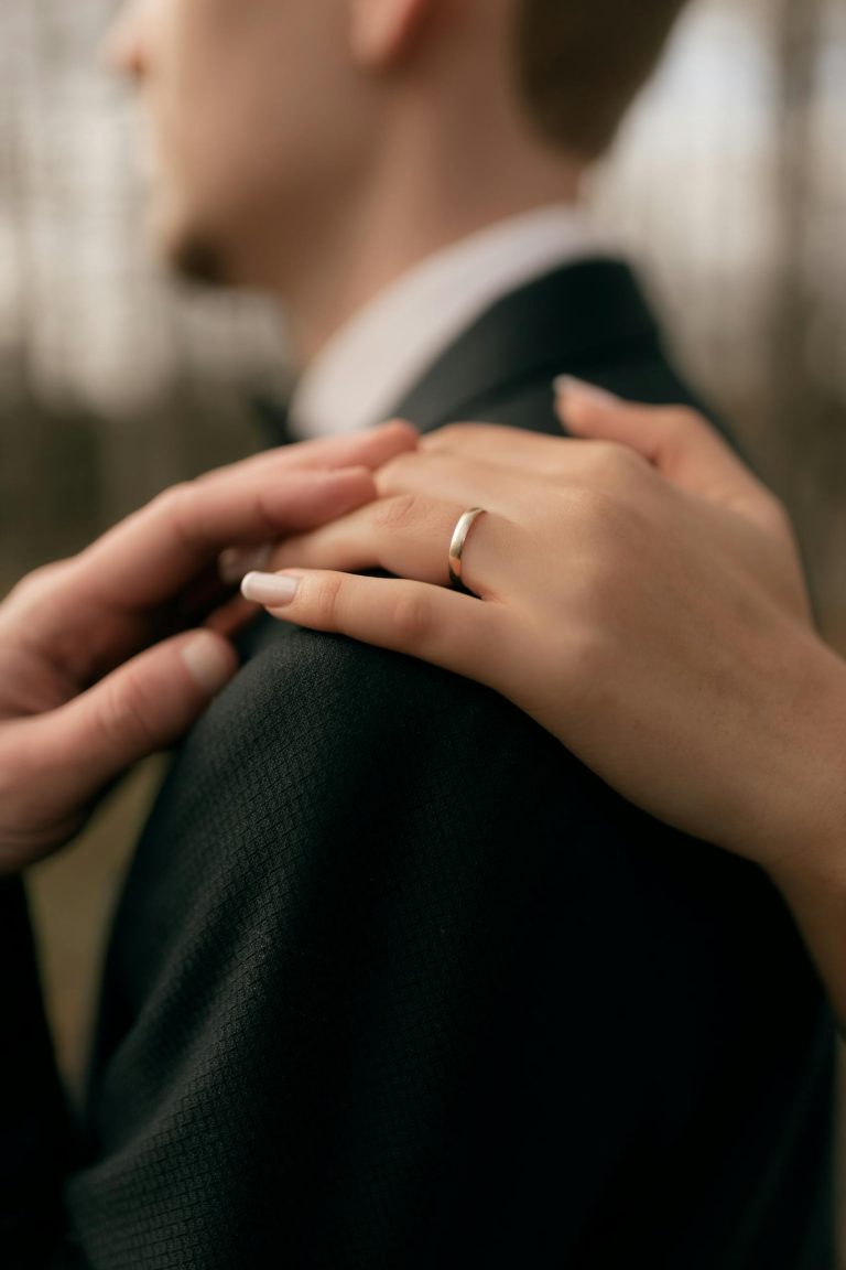 Close-up of hands on a shoulder, highlighting a silver ring. Warm and intimate moment outdoors.